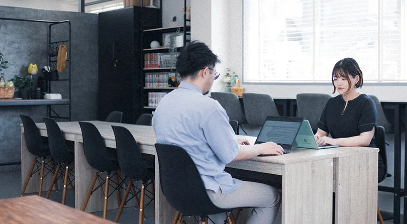 明るいオフィスで男女が向かい合い、それぞれノートパソコンで作業している写真
